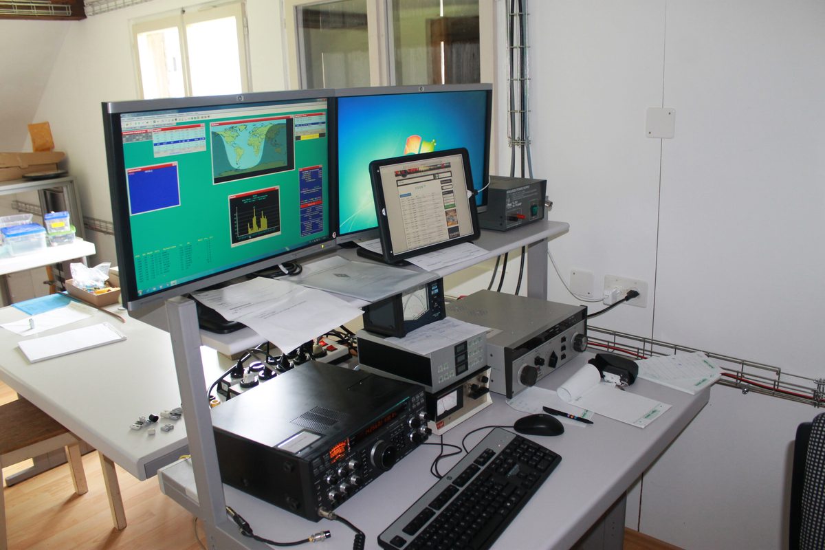 Arbeitsplatz einer Kurzwellenstation: zwei Monitore mit Logbuch und Weltkarte, davor Transceiver, Netzteil und Tastatur auf einem hellen Schreibtisch
