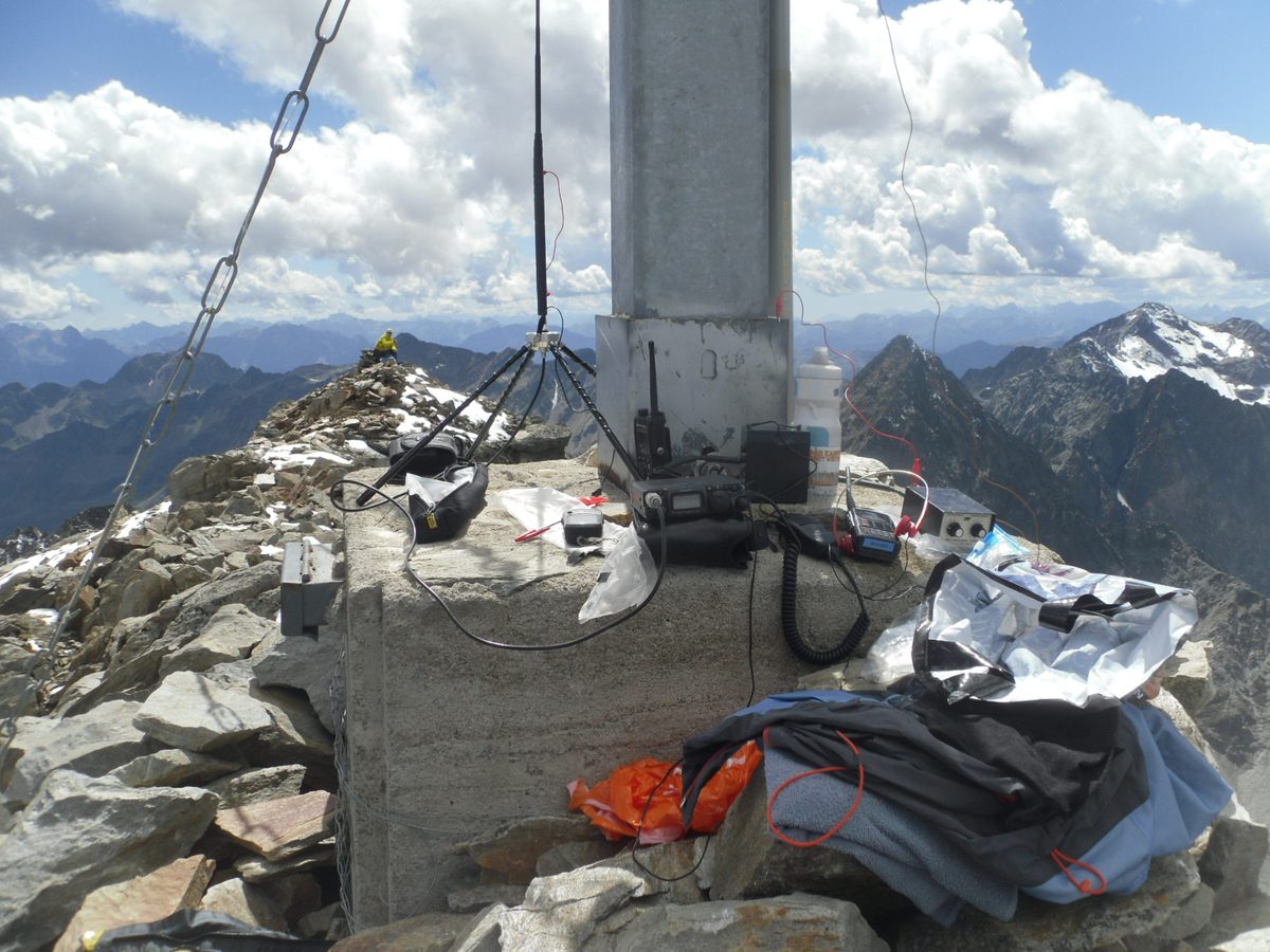 Gipfelkreuz-Sockel mit Handfunkgerät, kleinem Kurzwellengerät, Kabeln und Rucksack, dahinter Bergketten mit Schneeresten unter Wolken