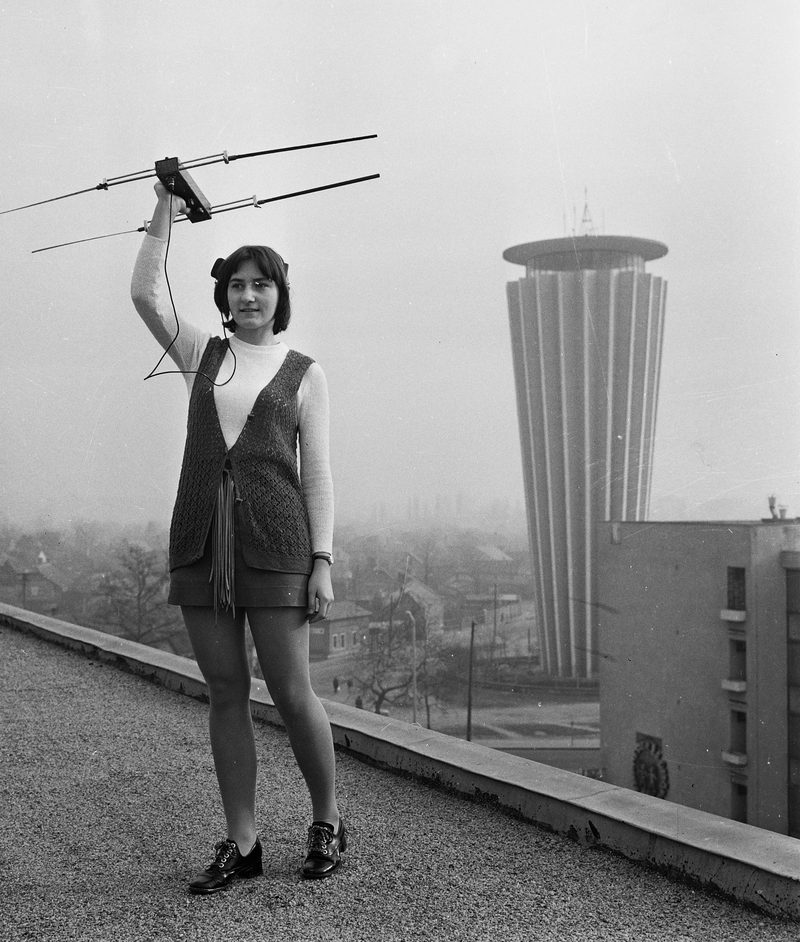 Schwarzweißfoto von 1973: Eine junge Frau steht auf einem Flachdach und hält eine kleine Richtantenne in die Höhe, im Hintergrund ein Wasserturm