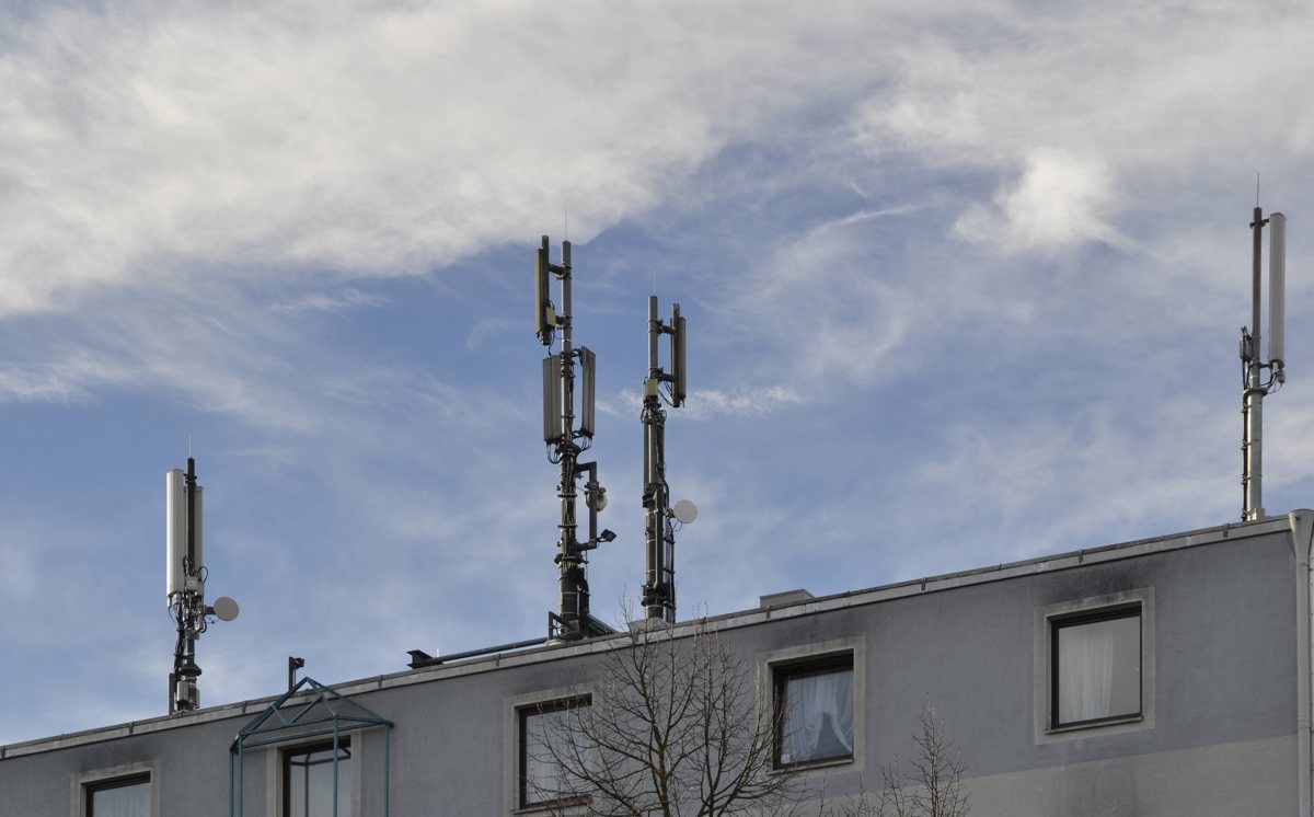 Vier schlanke Antennenmasten mit grauen Sektorantennen und kleinen Richtfunkschüsseln auf dem Flachdach eines Wohnblocks vor blauem Himmel