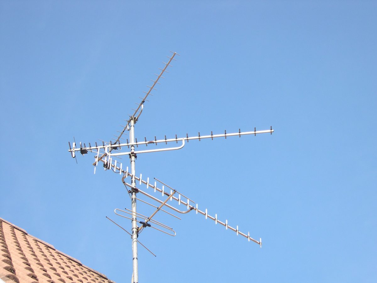 Drei Yagi-Antennen mit vielen kurzen Elementen an einem Mast auf einem Ziegeldach vor blauem Himmel, in verschiedene Richtungen ausgerichtet