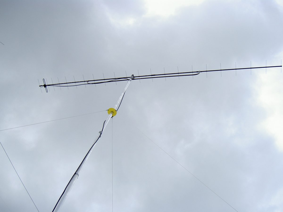 Sehr lange Yagi-Antenne mit vielen kurzen Elementen auf einem Tragrohr, schräg vor grauem Himmel auf einem Mast mit Abspannseilen