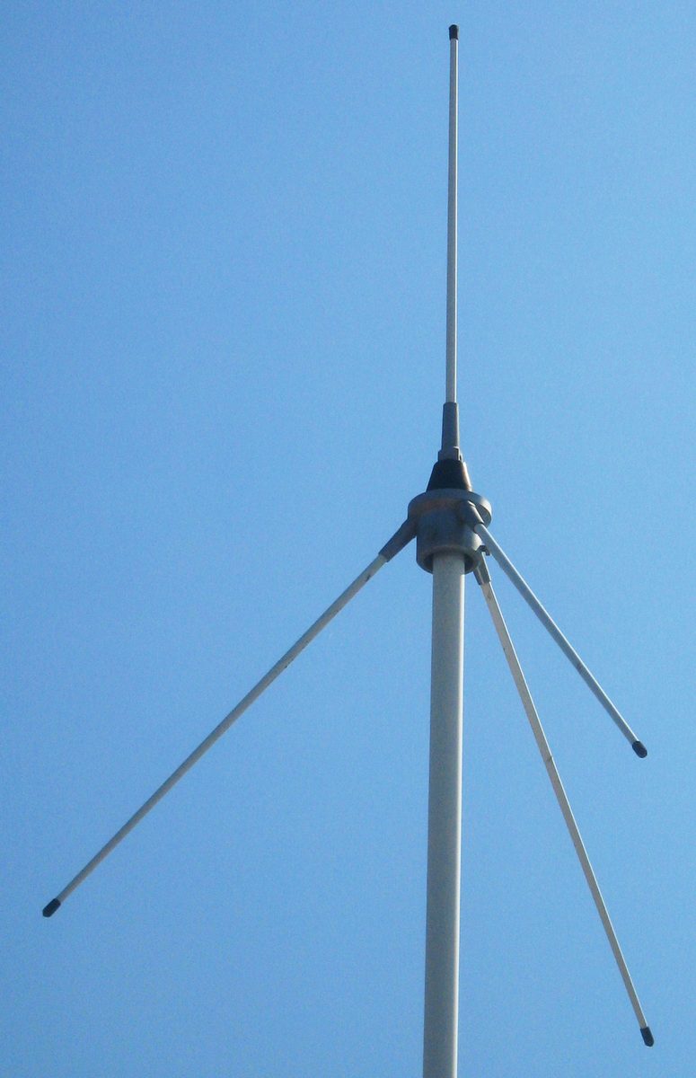 Senkrechter Stabstrahler auf einem Mastrohr vor blauem Himmel, am Fußpunkt vier schräg nach unten abstehende Radiale