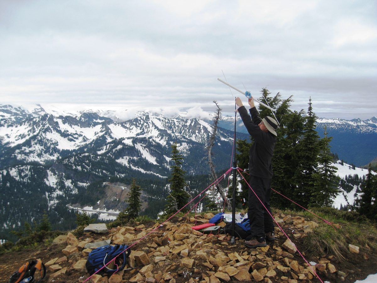 Ein Mann richtet auf einem steinigen Gipfel eine kleine Yagi-Antenne an einem Stativmast aus, im Hintergrund schneebedeckte Berge unter Wolken