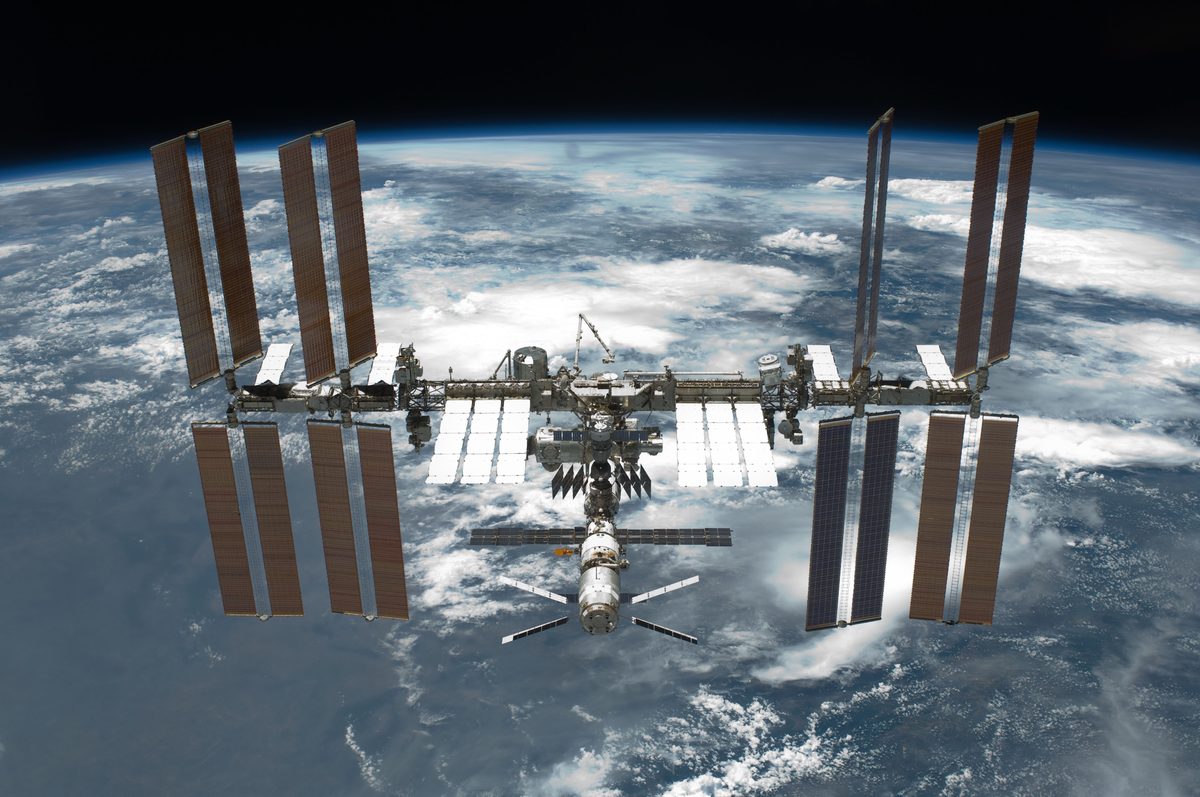 Die ISS mit acht großen braunen Solarflügeln vor der blau-weißen Erde, in der Mitte die Druckmodule, unten das russische Segment mit angedocktem Raumschiff