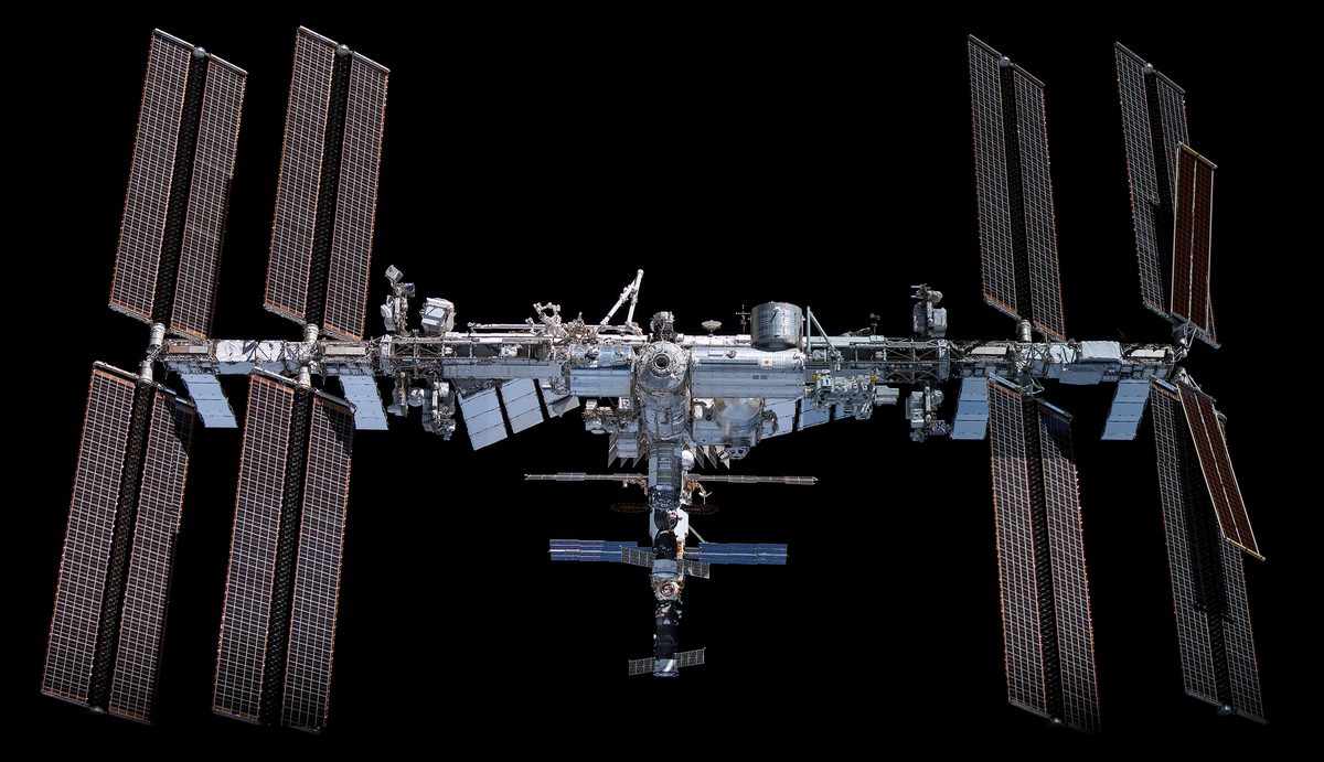 Die ISS vor schwarzem Himmel in voller Breite: links und rechts je vier große Solarflügel, rechts außen zwei kleinere schräg sitzende Flächen, in der Mitte die Modulkette