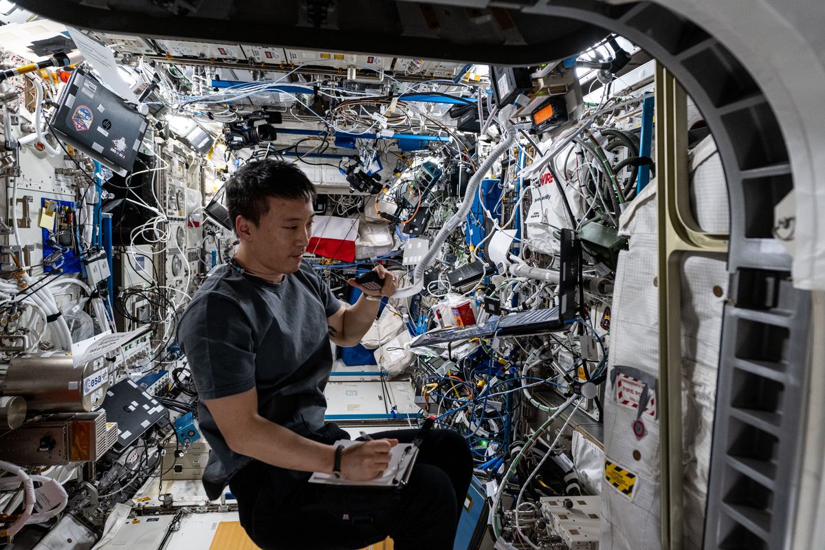 Astronaut Jonny Kim im vollgepackten Labor Columbus hält ein Mikrofon ans Ohr und schreibt auf einen Notizblock, um ihn herum Kabel, Geräte und Laptops