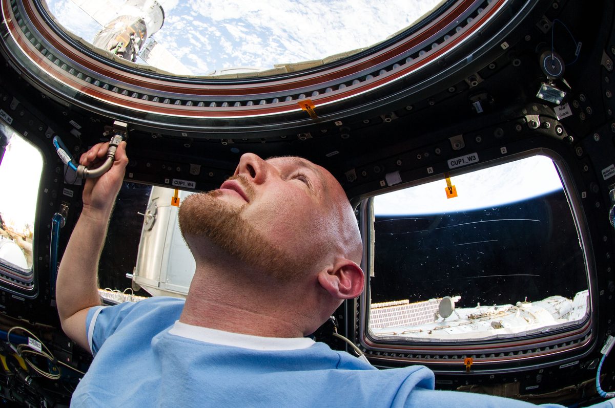 Alexander Gerst in hellblauem Shirt blickt nach oben durch die Fenster der Cupola, draußen die wolkenbedeckte Erde und Teile der Station