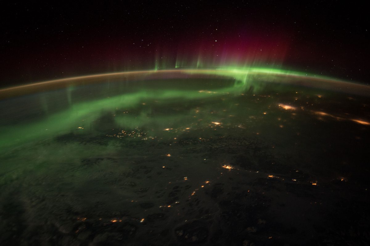 Blick aus der ISS auf die nächtliche Erde: grün und rot leuchtendes Polarlicht über dem Horizont, darunter die orangen Lichtpunkte von Städten