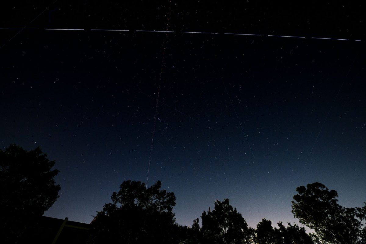 Nachthimmel über Bäumen mit der unterbrochenen hellen Spur der ISS, dünnen Satellitenspuren, kurzen Sternschnuppen und der roten Punktlinie eines Flugzeugs