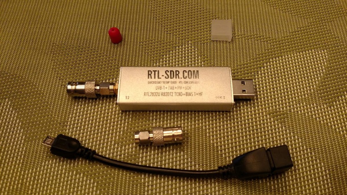 Silberner RTL-SDR-Stick mit USB-Stecker, daneben ein SMA-auf-BNC-Adapter und ein kurzes Micro-USB-Kabel auf einer Unterlage